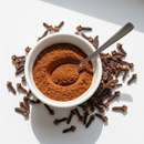 Ground cloves spice powder in a white ceramic bowl with whole clove buds on white background